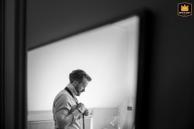 Artistic black and white profile shot of the groom’s reflection in a mirror, putting on his tie in the bedroom at Fawsley Hall.